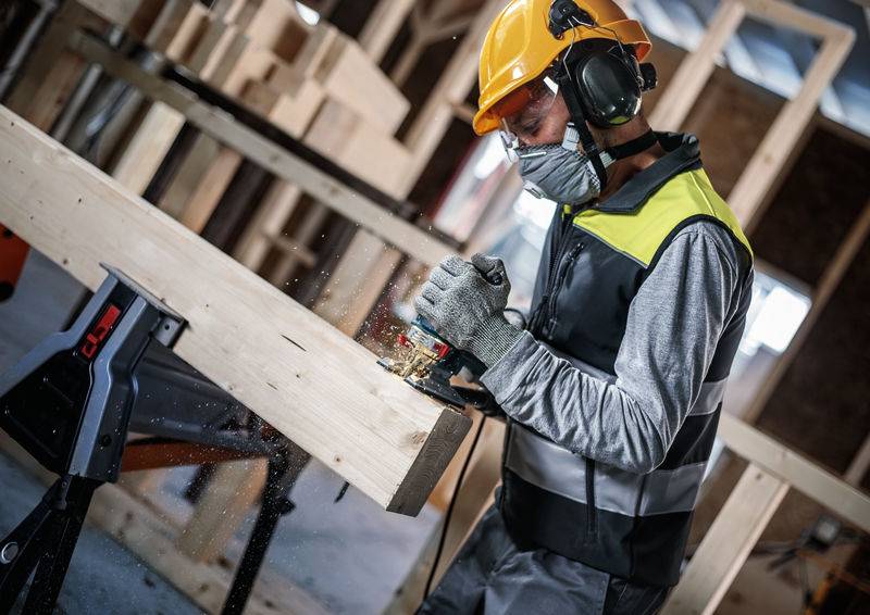 Ein Arbeiter mit Schutzhelm und Gehörschutz schleift ein Holzbrett. Funken fliegen. Hintergrund zeigt eine Baustelle.