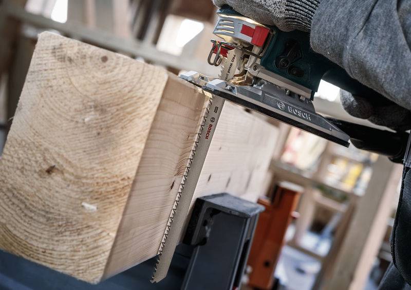 Eine Person benutzt eine elektrische Stichsäge, um ein großes Stück Holz zu schneiden. Es entsteht Sägemehl, das um das Holz verstreut ist.
