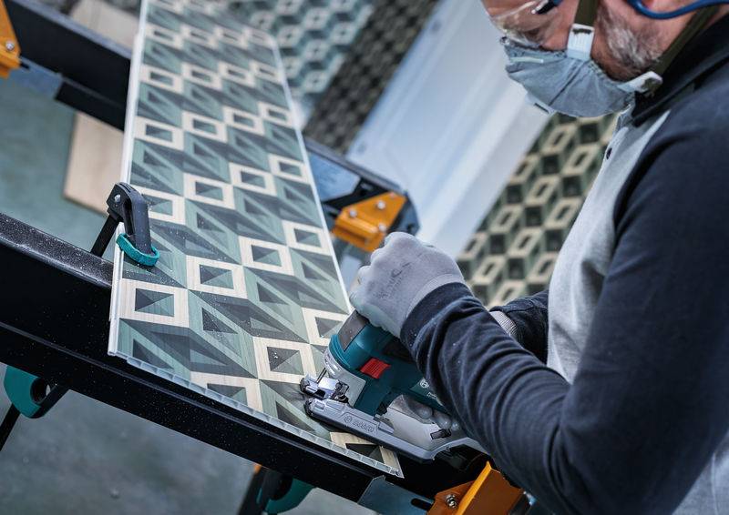 Eine Person schneidet ein geometrisch gemustertes Holzbrett mit einer elektrischen Säge. Schutzbrille und Maske werden getragen.