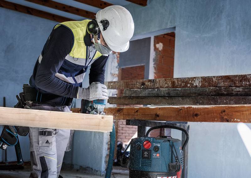 Ein Arbeiter mit Schutzhelm und -brille verwendet eine elektrische Handsäge, um ein Holzbrett in einer Werkstatt zu schneiden.