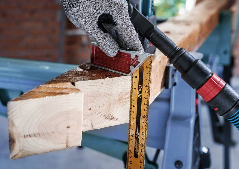 Person mit Handschuh misst ein Holzbrett mit einem Winkelmesser und Lineal, während ein Bohrwerkzeug in der Nähe bereitsteht.