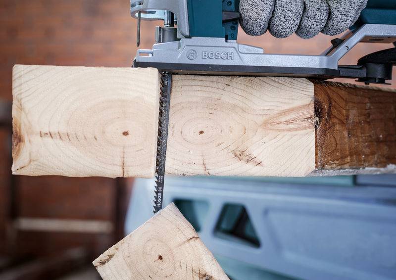 Ein Elektrowerkzeug sägt einen Holzbalken. Ein Stück Holz fällt ab. Hintergrund unscharf, Fokus auf das Sägeblatt und den Schnitt.