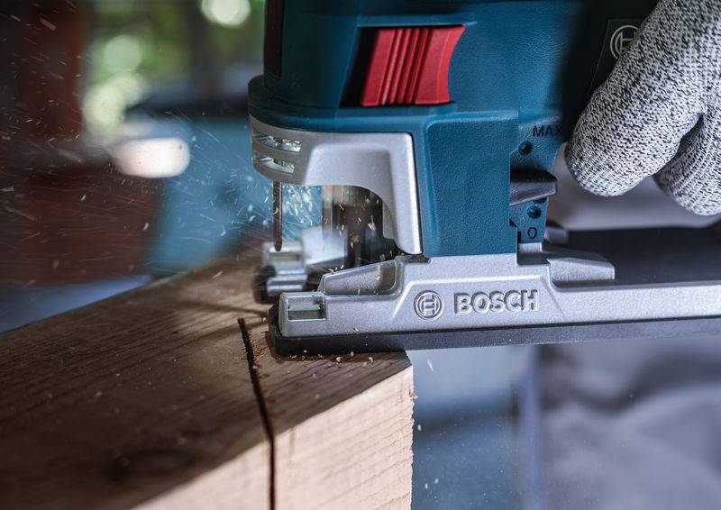 Eine Stichsäge schneidet präzise durch ein Holzbrett, während Sägespäne fliegen. Ein Handschuh hält das Holz stabil.