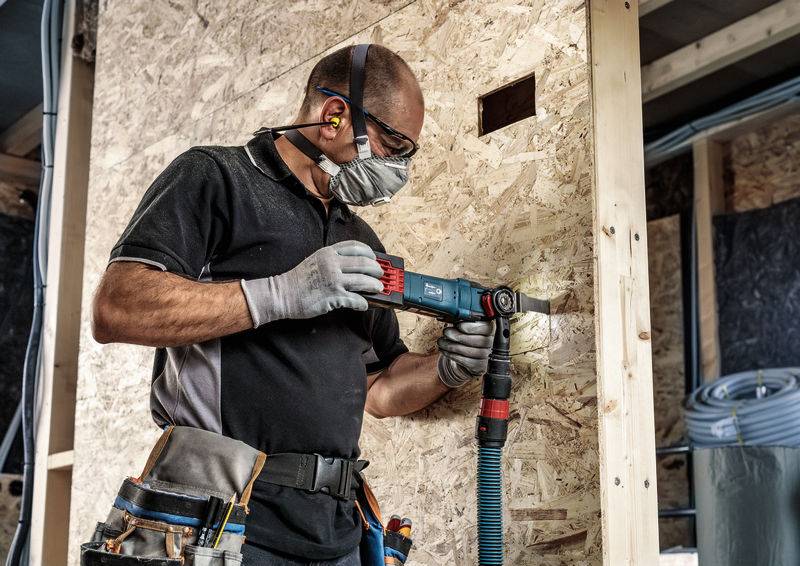 Ein Arbeiter bohrt mit Schutzbrille, Maske und Gehörschutz ein Loch in eine Holzplatte. Werkzeugkasten an der Hüfte befestigt.