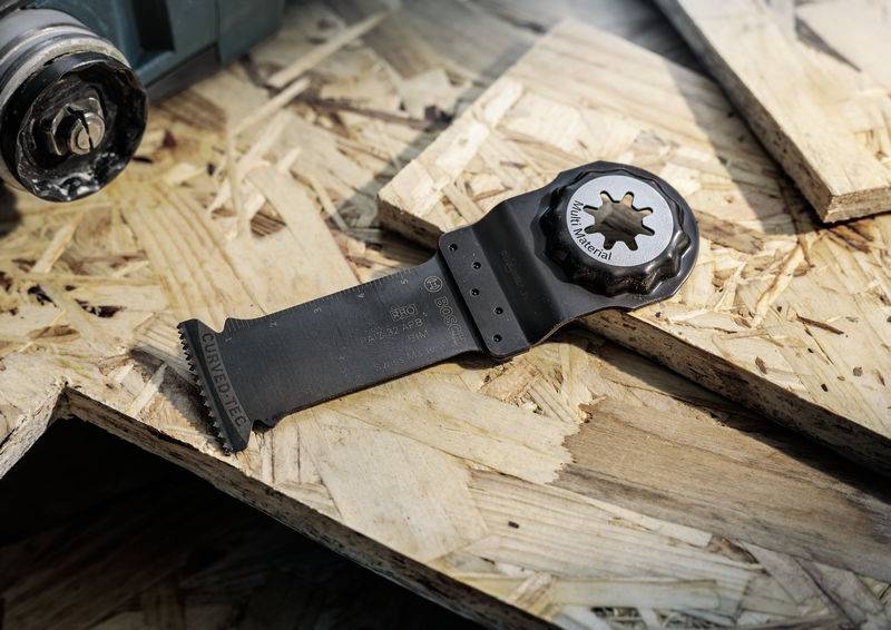 Ein schwarzes Sägeblatt für ein Multitool liegt auf einer Holzoberfläche. Im Hintergrund ist ein Teil eines Werkzeugs sichtbar.