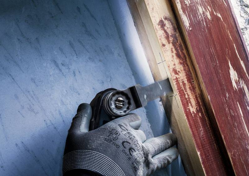 Hand mit Handschuh, die eine elektrische Säge benutzt, um ein Holzstück an einem Fensterrahmen zu schneiden. Renovierungsarbeit im Gange.