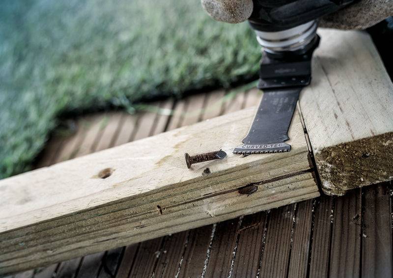 Elektrowerkzeug schneidet Holzbrett nahe einer verrosteten Schraube auf einem Holzboden. Im Hintergrund ist Kunstrasen zu sehen.