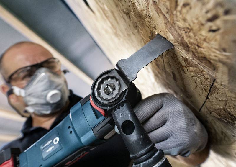 Ein Handwerker trägt Schutzbrille und Atemmaske, während er mit einem blauen Elektrowerkzeug an einer Holzplatte im Innenraum arbeitet.