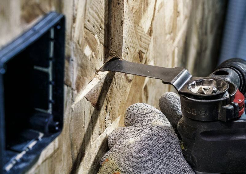 Eine Person in Handschuhen verwendet ein Multitool, um ein Quadrat in eine Holzplatte zu schneiden. Links ist eine leere Steckdosenöffnung.