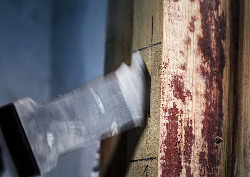 Ein eingehauener Handbeil nähert sich einem Holzbalken, während es im Holz Spuren hinterlässt, was auf Tischlerarbeit hindeutet.