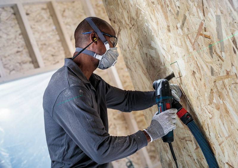 Ein Arbeiter trägt Schutzbrille und Maske, bohrt mit einer Bohrmaschine in eine OSB-Holzplatte. Sicherheitsvorkehrungen im Vordergrund.