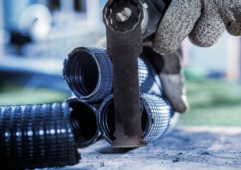 Hand mit Arbeitshandschuh hält ein oszillierendes Werkzeug, das an Wellenstutzen aus Metall arbeitet. Im Hintergrund unscharf.
