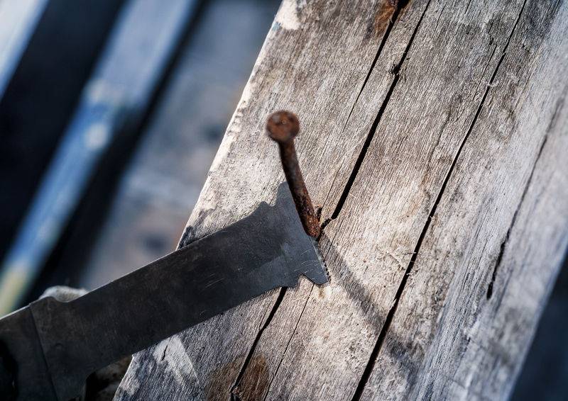 Eine verrostete Nagelspitze ragt aus einem alten Holzbalken, daneben eine Säge mit einer abgebrochenen Klinge.