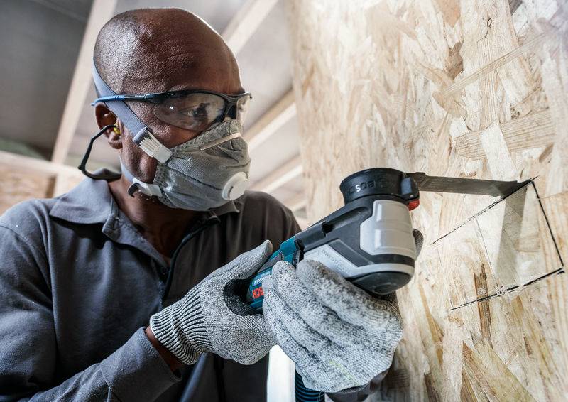 Ein Arbeiter sägt mit einer elektrischen Säge eine rechteckige Öffnung in eine Sperrholzplatte, trägt Schutzbrille und Atemmaske.