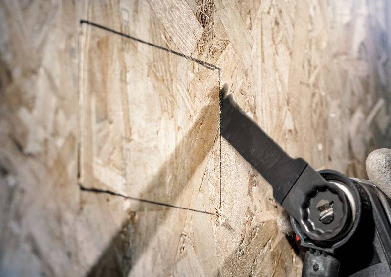 Eine Hand führt eine Elektrosäge, schneidet präzise ein Rechteck in eine Holzplatte.