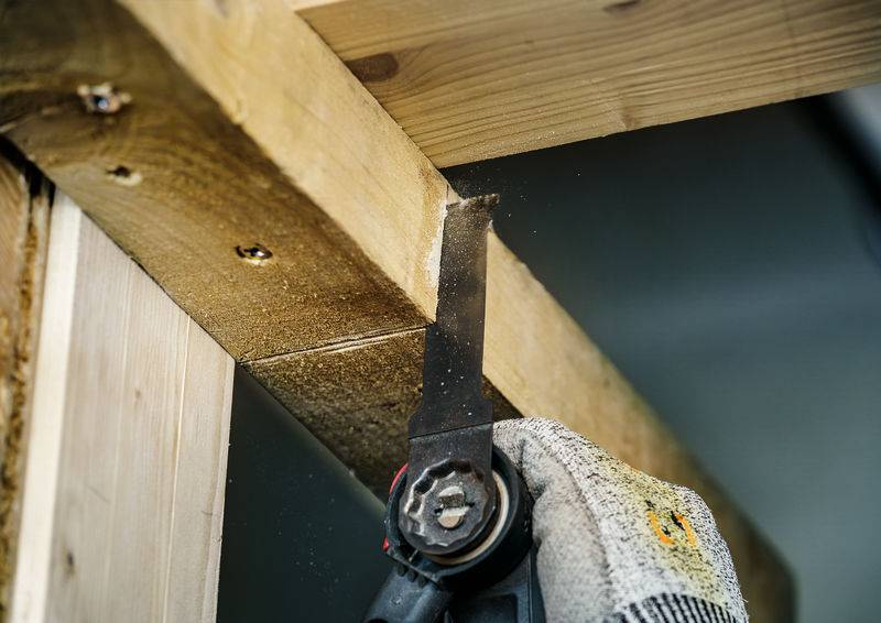 Arbeiter sägt Holz mit einer elektrischen Säge. Eine behandschuhte Hand hält die Säge an einem hölzernen Balken in einem Gebäude.