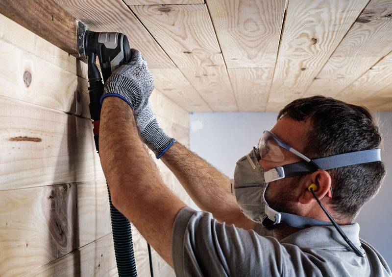Ein Mann trägt Schutzkleidung und schleift mit einem Elektrowerkzeug Holzpaneele an der Decke.