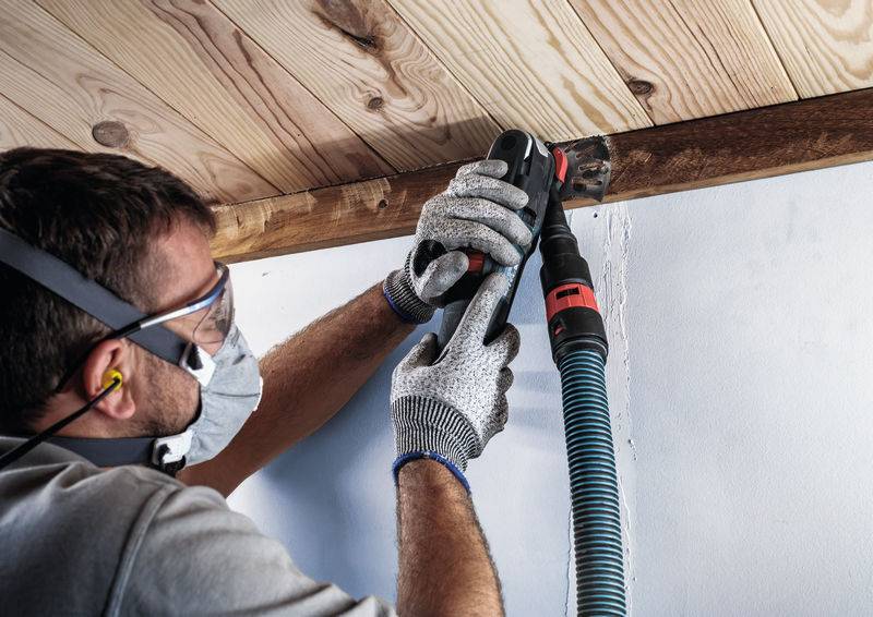 Eine Person mit Schutzbrille und Maske, die mit einem elektrischen Schleifgerät eine Holzkante an einer Decke bearbeitet.