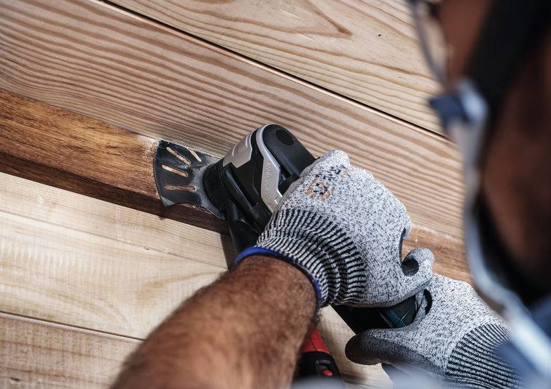 Eine Person sägt Holz mit einem elektrischen Werkzeug, trägt dabei graue Handschuhe und Schutzbrille. Fokus liegt auf der Arbeitssituation.