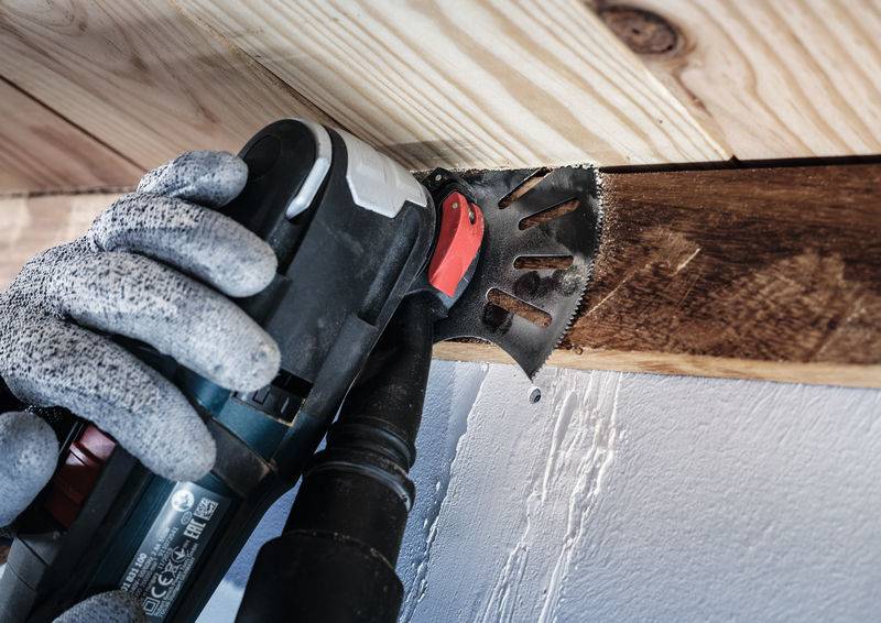 Person mit grauen Handschuhen benutzt ein elektrisches Multitool, um Holzplanken an der Decke bündig zu schneiden.