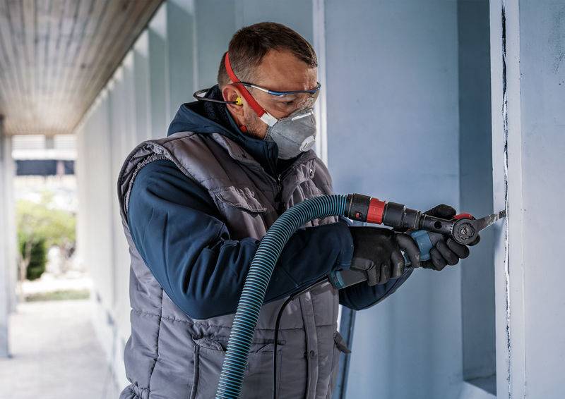 Eine Person arbeitet mit Schutzbrille, Maske und Gehörschutz an einer Wand. Sie verwendet ein handgehaltenes Werkzeug mit Schlauch.