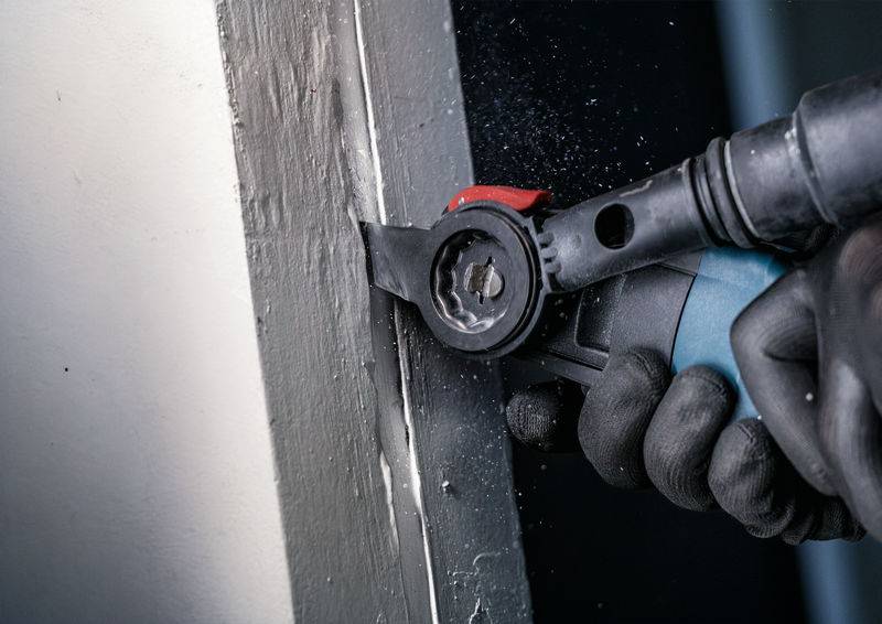 Person mit Schutzhandschuhen hält ein Multitool und schneidet präzise entlang einer Ecke einer Wand.