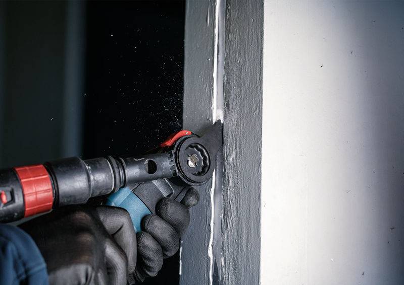 Eine Person schneidet mit einem oszillierenden Werkzeug Dichtungsmaterial aus einer weißen Wandfuge. Sie trägt schwarze Handschuhe.