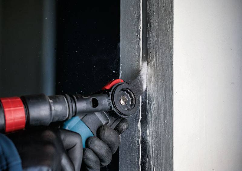 Ein Arbeiter trägt Handschuhe und benutzt ein Elektrowerkzeug, um einen grauen Türrahmen zu schleifen.