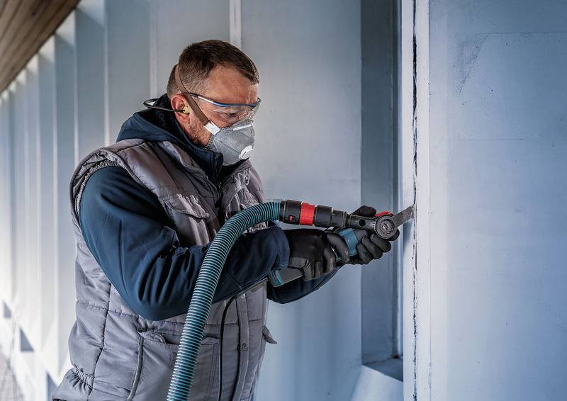 Eine Person trägt Schutzkleidung und benutzt ein Elektrowerkzeug, um in einer Betonwand zu arbeiten. Schutzbrille und Maske sind sichtbar.