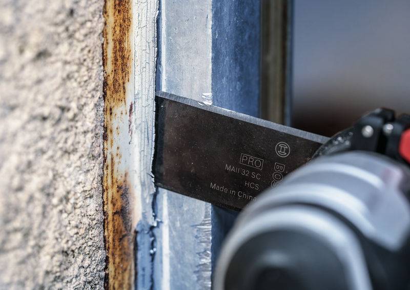 Ein Metallwerkzeug in der Nahaufnahme löst Rost von einem metallischen Rahmen neben einer Betonwand.