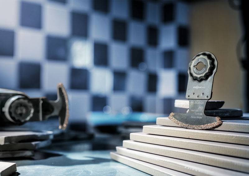 Ein oszillierendes Sägeblatt steht auf gestapelten Fliesen, im Hintergrund ist eine verschwommene Kachelwand sichtbar.