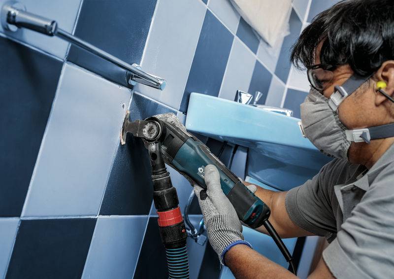 Eine Person trägt Schutzmaske und Gehörschutz, verwendet eine elektrische Schleifmaschine, um Fliesen in einem Badezimmer zu bearbeiten.