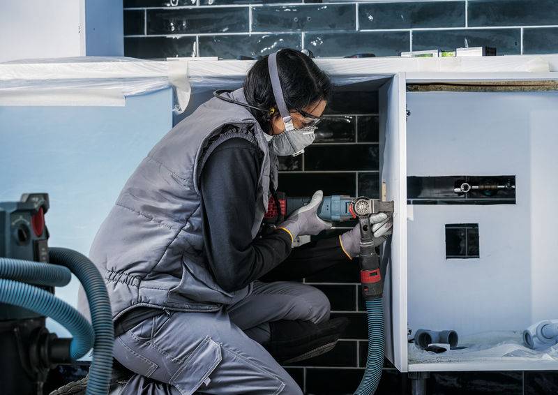 Eine Person arbeitet mit einer Bohrmaschine in einer Küche. Sie trägt Schutzkleidung und eine Maske, um Sicherheitsvorkehrungen einzuhalten.