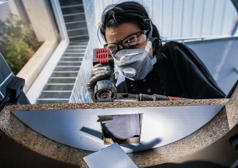 Eine Person trägt Schutzbrille und Maske und arbeitet konzentriert mit einer Bohrmaschine an einer Holzplatte in einem lichtdurchfluteten Raum.