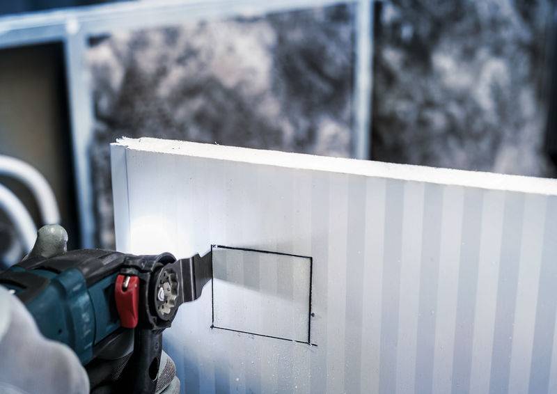 Eine Person schneidet mit einem elektrischen Werkzeug ein Rechteck in eine Gipskartonplatte.