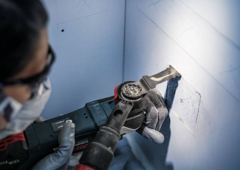 Eine Person bearbeitet mit einem Elektrowerkzeug eine Wand. Schutzbrille und Maske werden getragen, um Sicherheit beim Arbeiten zu gewährleisten.