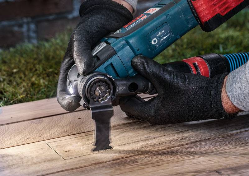Eine Person in Arbeitshandschuhen schneidet mit einem Elektrowerkzeug Holzplanken. Die Szene zeigt handwerkliche Tätigkeit im Freien.