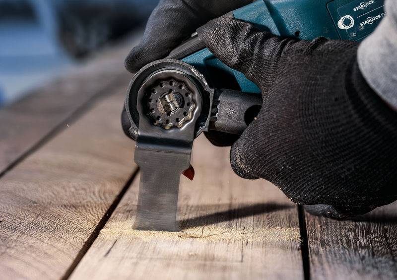 Ein Handwerker trägt schwarze Handschuhe und benutzt ein Elektrowerkzeug, um Holz zu sägen. Feiner Holzstaub wird aufgewirbelt.