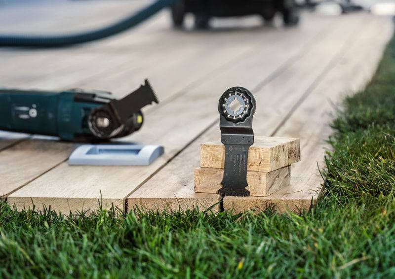 Eine elektrische Säge mit einem Holzschneideblatt liegt auf einer Holzterrasse, daneben geschnittene Holzstücke und Rasen.