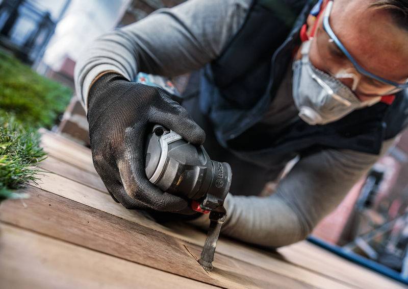 Person mit Schutzbrille und Maske arbeitet mit einem Elektrowerkzeug an Holzplanken im Freien.