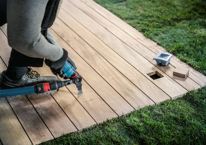 Eine Person installiert Holzdielen im Garten, verwendet ein Elektrowerkzeug neben vorbereiteten Aussparungen.