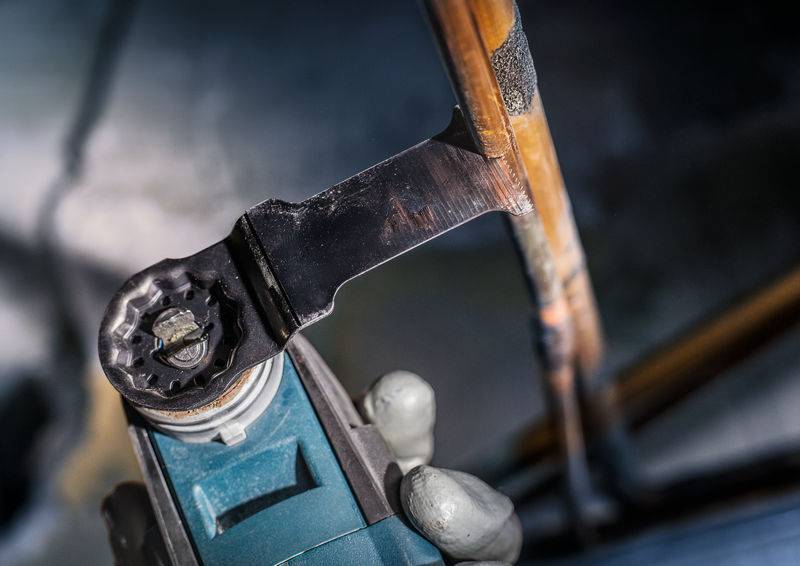 Eine Person schneidet ein Kupferrohr mit einem Elektrowerkzeug. Die Umgebung deutet auf eine handwerkliche Tätigkeit oder Reparatur hin.