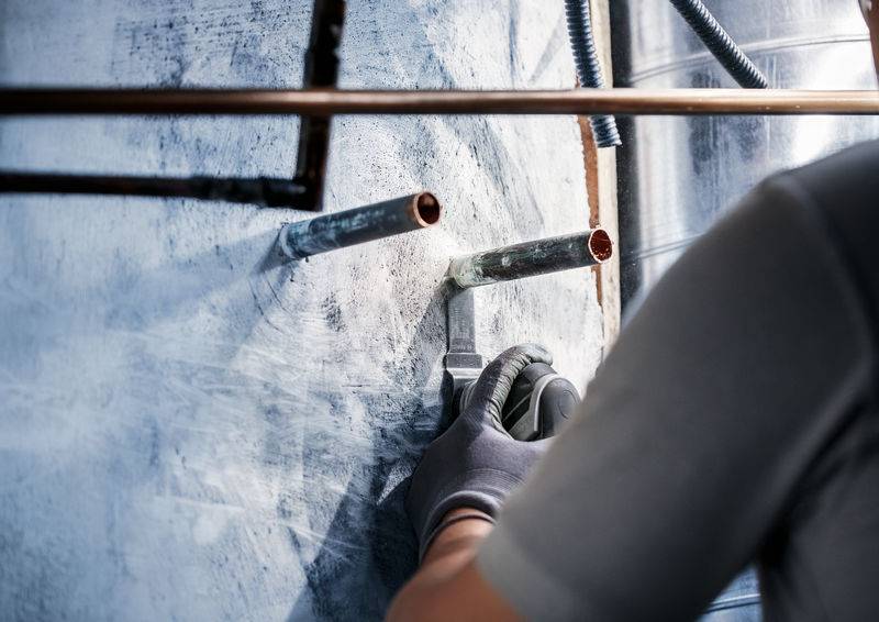 Ein Installateur arbeitet an einer Wand, schneidet Rohrleitungen. Er trägt Arbeitshandschuhe und nutzt ein Werkzeug, um Präzision zu gewährleisten.