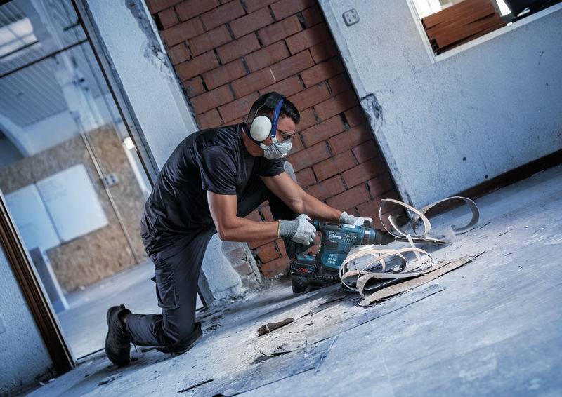 Arbeiter mit Gehörschutz und Schutzbrille bearbeitet mit einem Elektrowerkzeug einen Boden in einem im Bau befindlichen Gebäude.