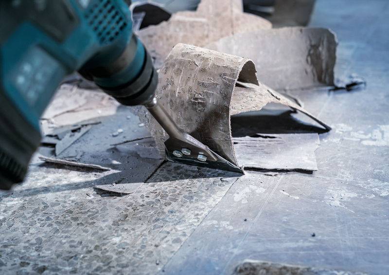 Ein Elektrowerkzeug entfernt Fliesenreste von einem Boden. Der Bohrer hebt brüchiges Material und Schmutz in einer Baustelle ab.