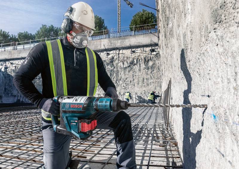 Arbeiter mit Schutzkleidung und Bohrmaschine arbeitet an Betonwand auf Baustelle. Im Hintergrund Baustellenstruktur und blauer Himmel.