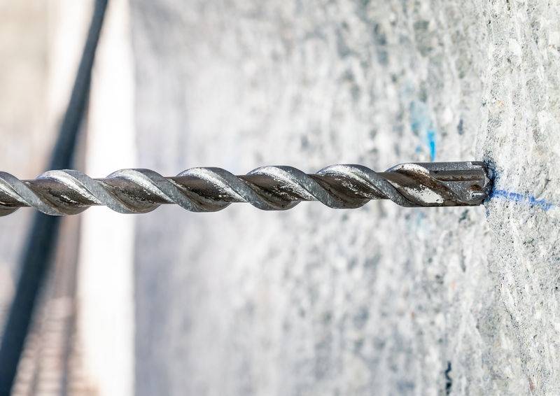 Ein metallischer Bohrer dringt in eine graue Betonwand ein, erzeugt feinen Staub und zeigt einen Punkt der Präzision in handwerklicher Arbeit.