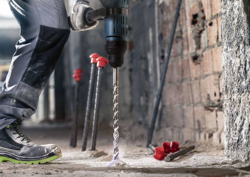 Person bohrt mit einer elektrischen Bohrmaschine in Betonboden. Im Hintergrund sind Metallstäbe und rote Markierungen an der Wand sichtbar.