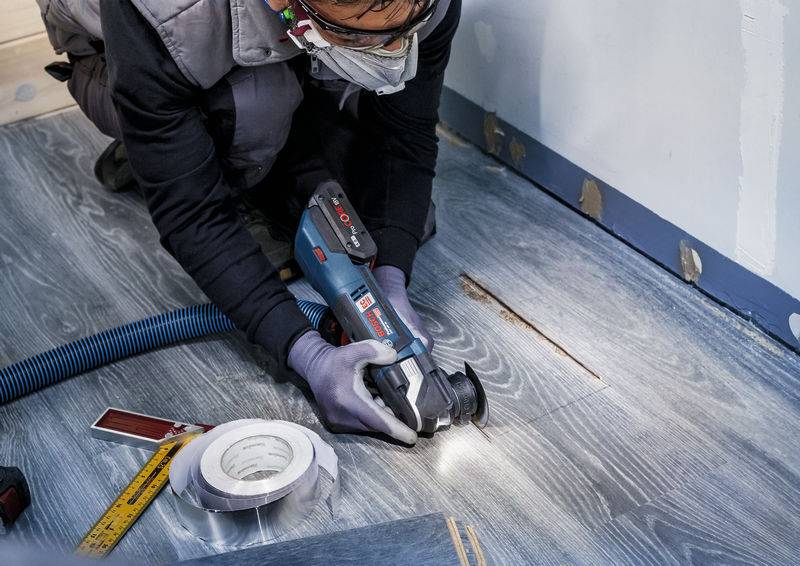 Person, die Schutzkleidung trägt, arbeitet mit einem Elektrowerkzeug an einem Holzboden. Ein Maßband und Klebeband liegen daneben.