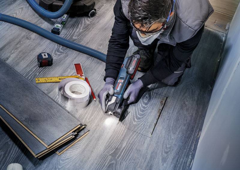 Eine Person arbeitet mit einem Elektrowerkzeug an Holzfußbodenplatten, umgeben von Maßband, Klebeband und Brettern.